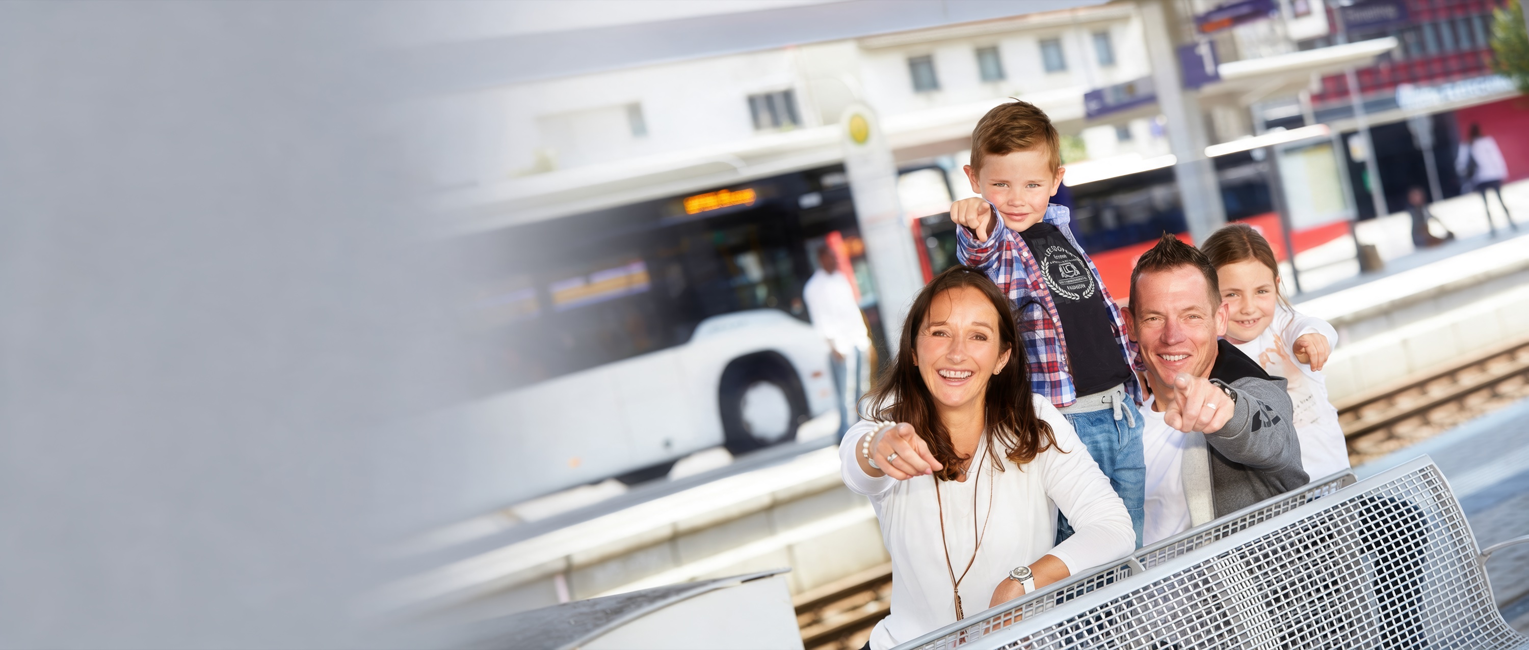 Lachende Familie auf einer Bank am Bahnsteig zeigt nach vorn, im Hintergrund fährt ein Bus