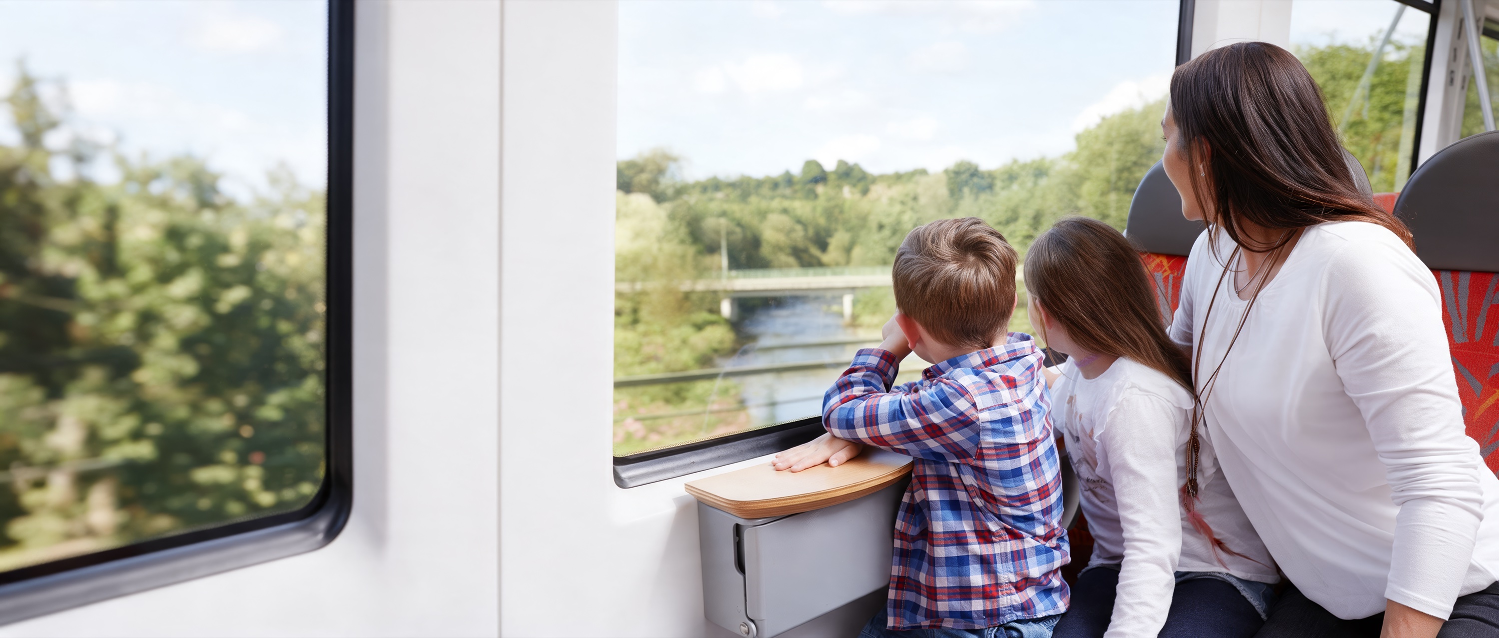 Mutter mit zwei Kindern im Zug, sie schauen aus dem Fenster auf einen Fluss mit Brücke